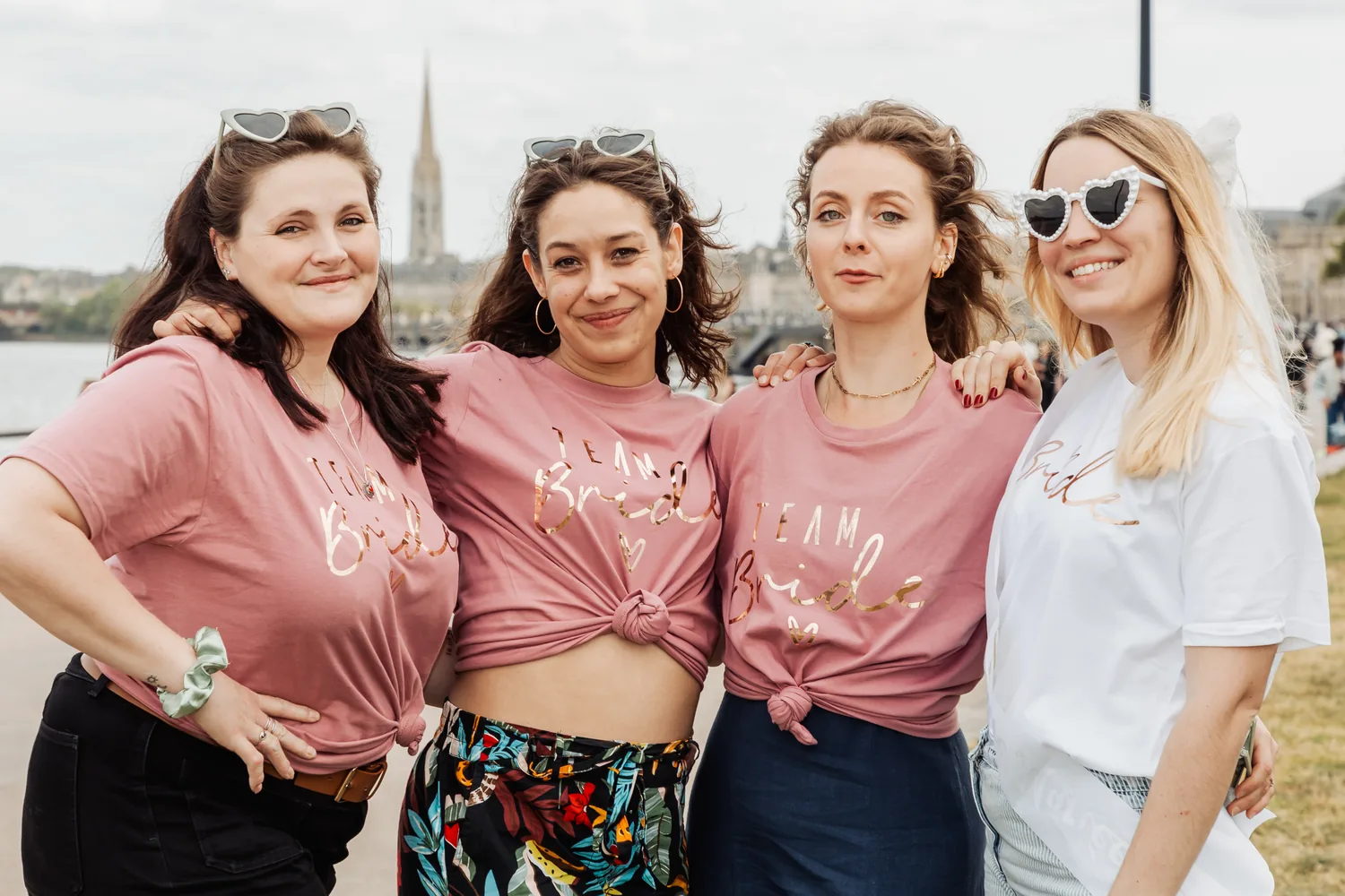 Quatre copines serrées épaule contre épaule au bord du fleuve, t-shirts Team Bride et lunettes en forme de cœur, EVJF à Bordeaux