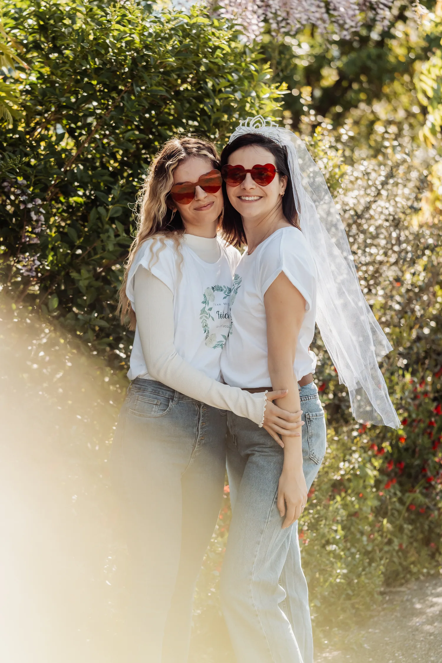 Deux amies enlacées dans un jardin ensoleillé, lunettes en forme de cœur, la mariée en voile blanc, EVJF à Bordeaux