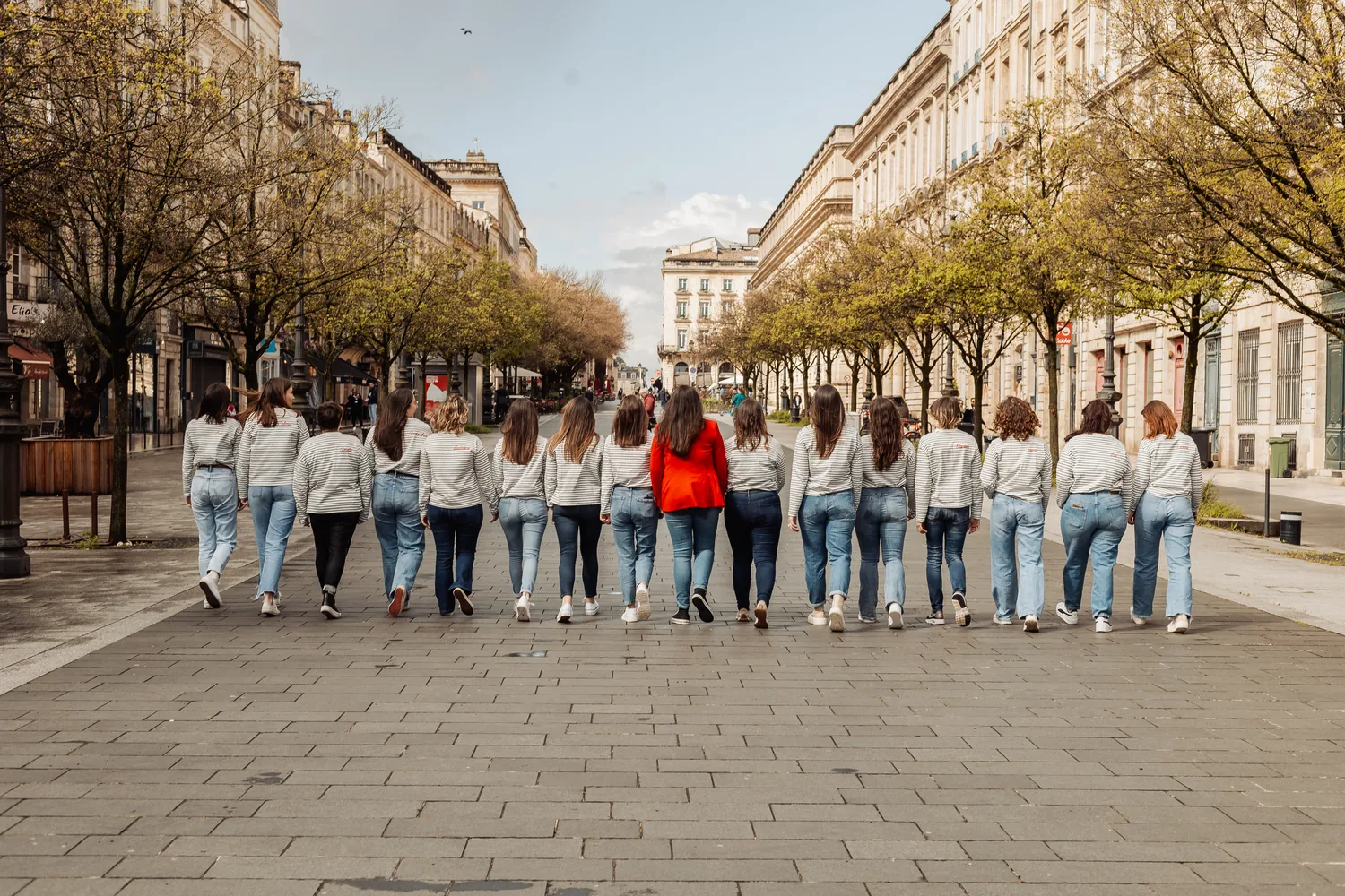Le groupe de copines en pulls assortis, de dos, remonte une large rue bordée d’arbres et de façades de pierre, EVJF à Bordeaux
