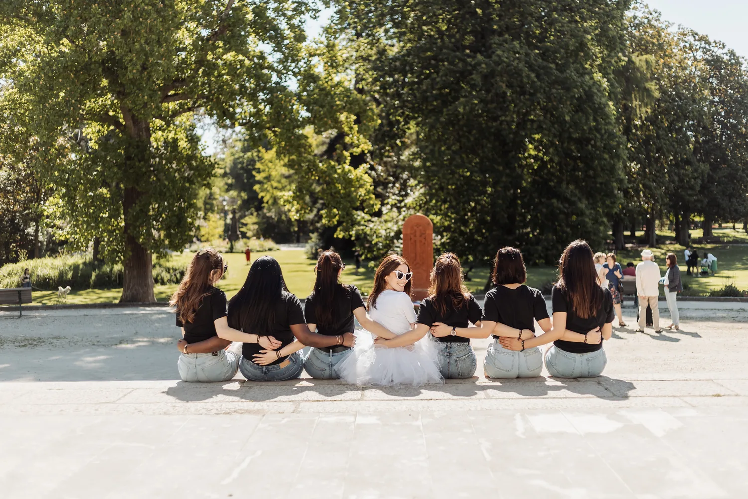 Sept copines assises côte à côte sur un muret, bras dessus bras dessous et de dos, face à un parc ensoleillé, EVJF à Bordeaux