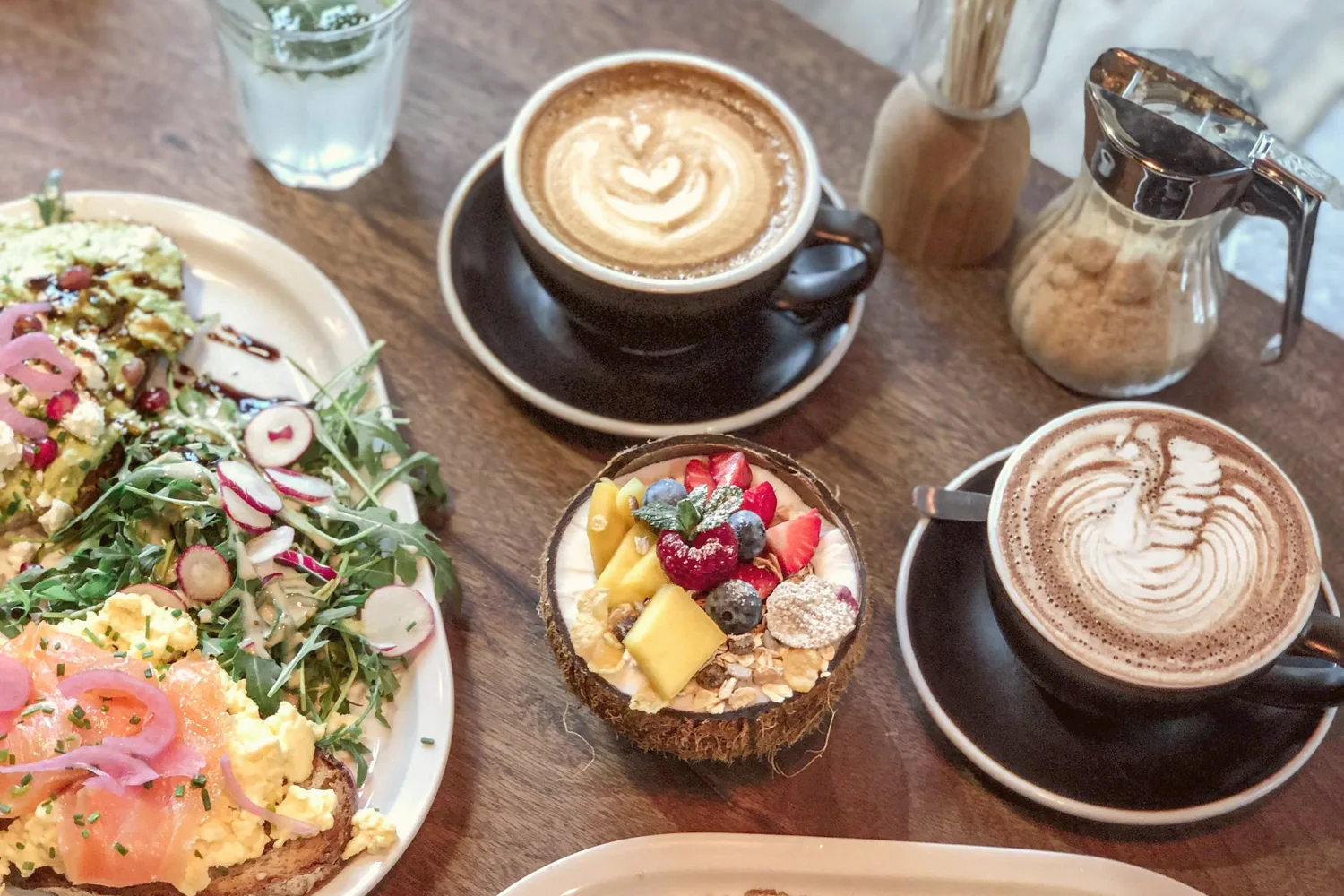 Table de brunch en bois : deux cafés crème, un bol de fruits frais dans une noix de coco et une assiette de tartines au saumon et à l'avocat