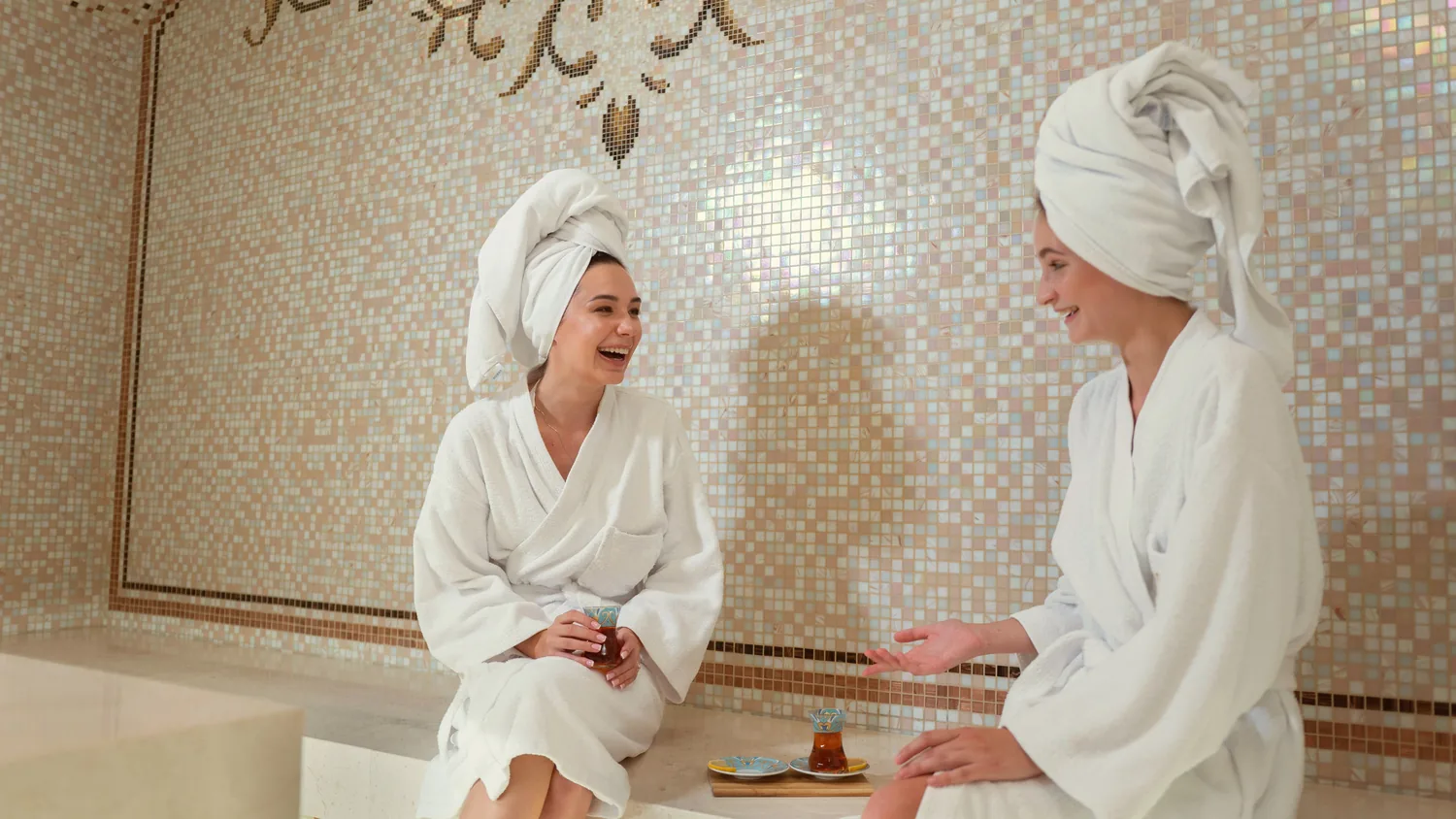 Deux amies en peignoir blanc, une serviette nouée sur la tête, rient autour d'un thé dans un hammam aux murs de mosaïque